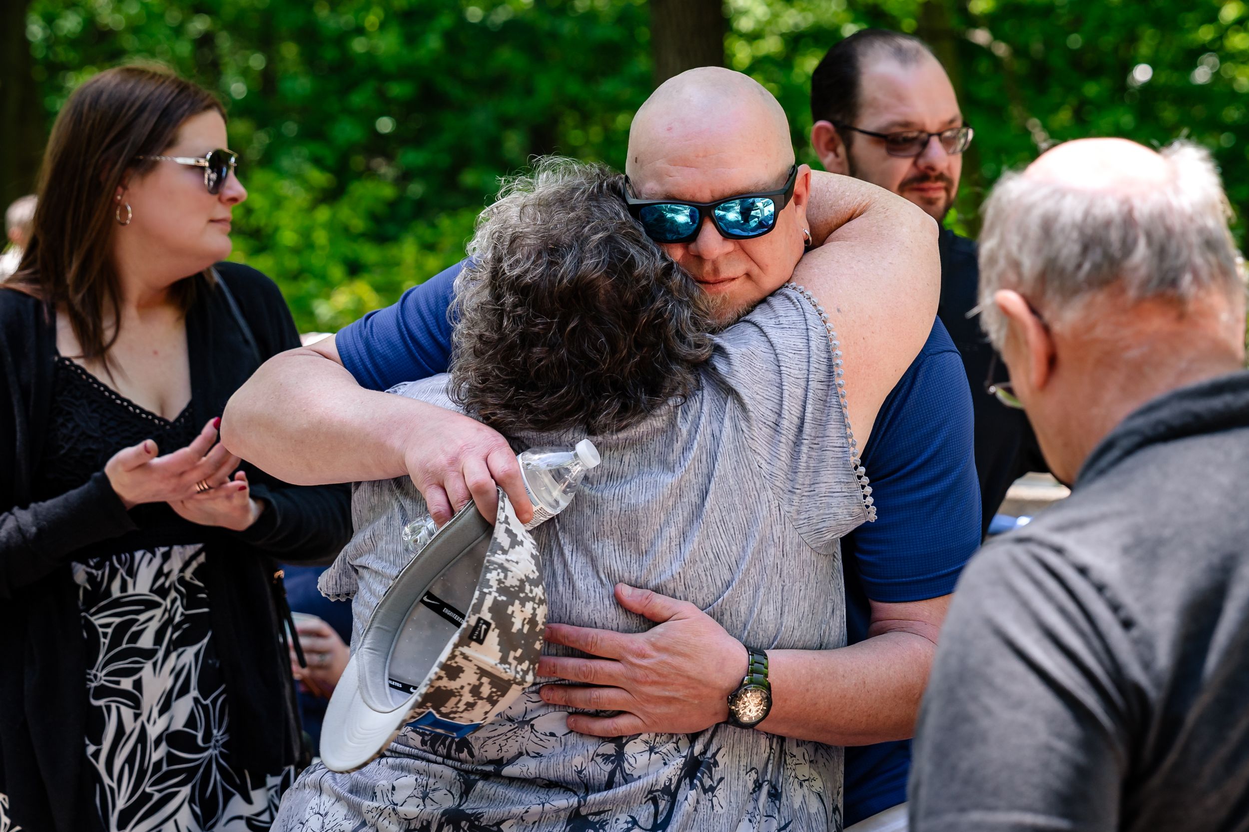Two people share an emotional hug outdoors surrounded by friends and family during a heartfelt gathering.