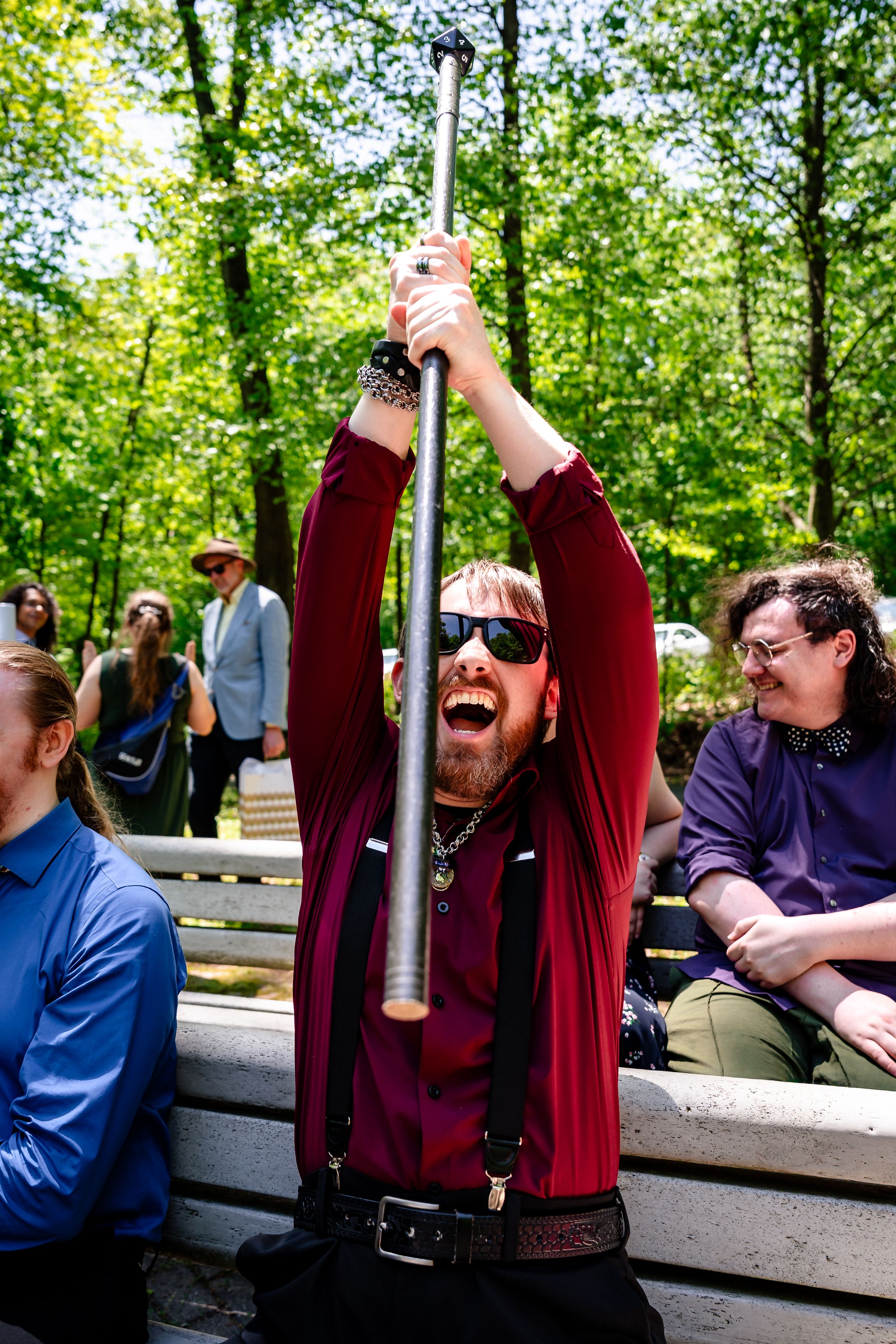Excited bearded man in red shirt raises metal pole overhead while sitting on bench at outdoor wedding.