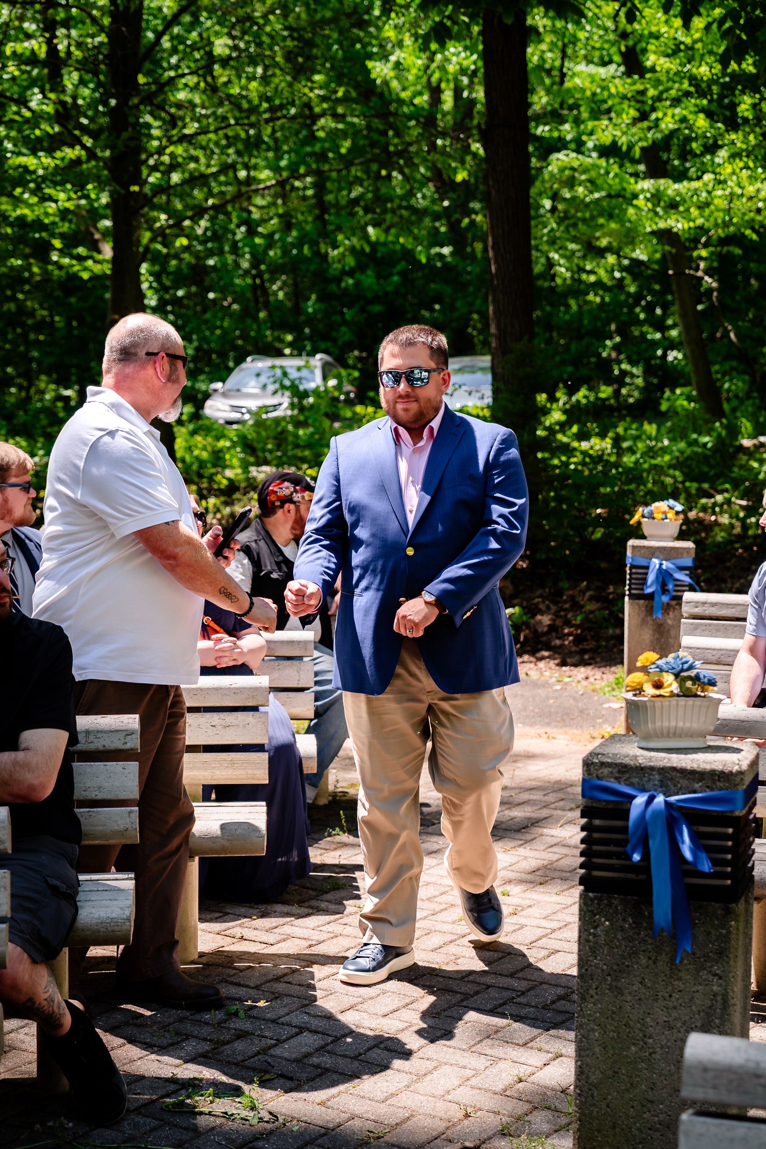 Man in navy blazer and khakis walks down outdoor wedding aisle surrounded by seated guests in wooded venue.