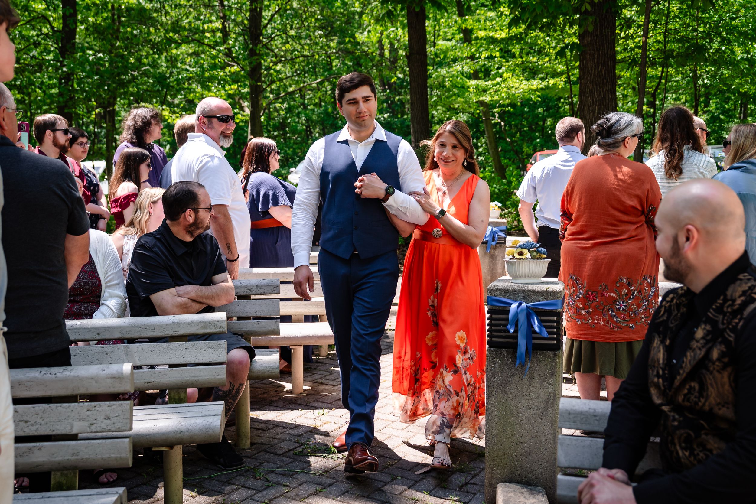 Groom in blue vest walks with woman in red dress through outdoor wedding reception among guests.