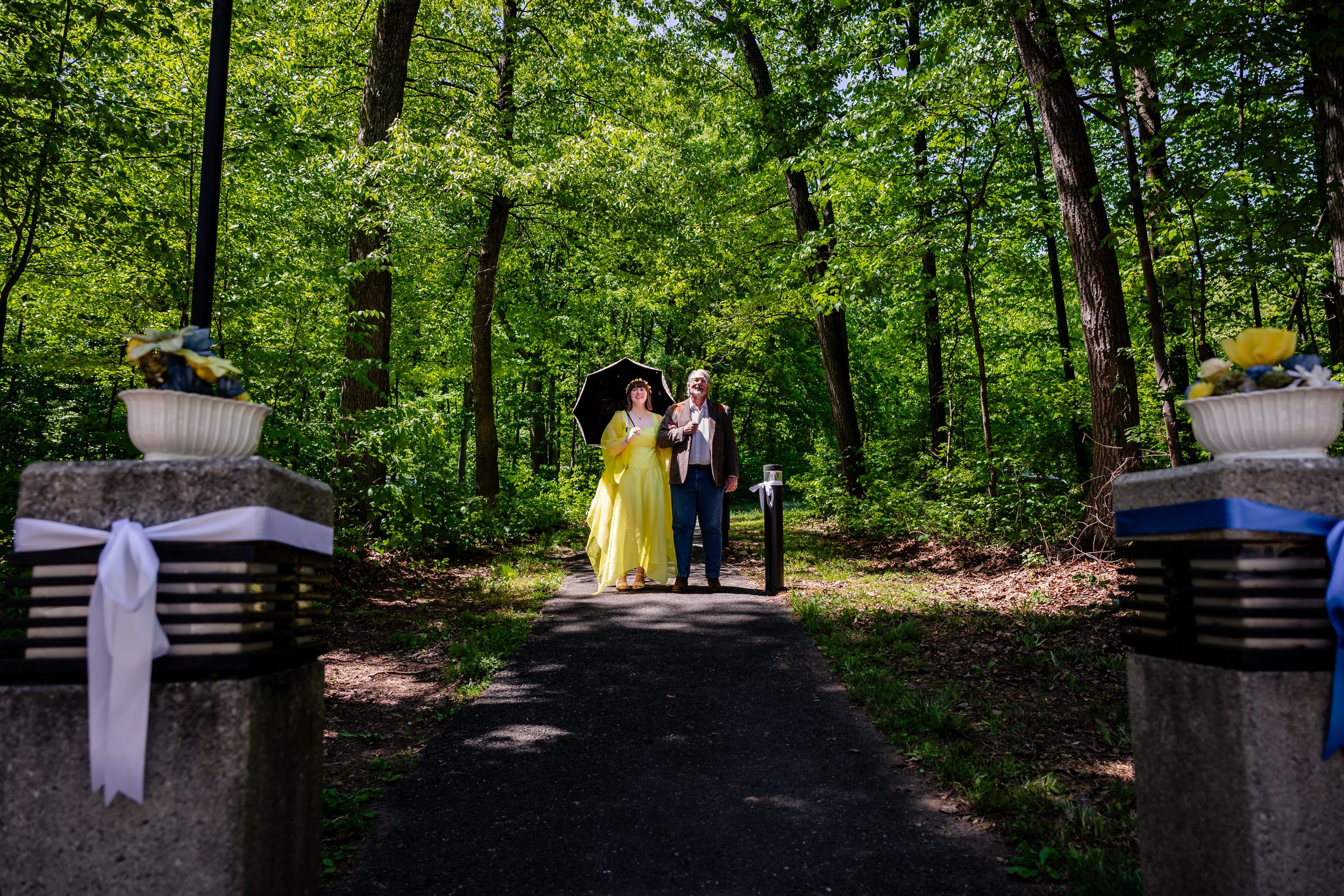 Bride in yellow gown and groom walk through stone gate entrance of lush green woodland wedding venue.