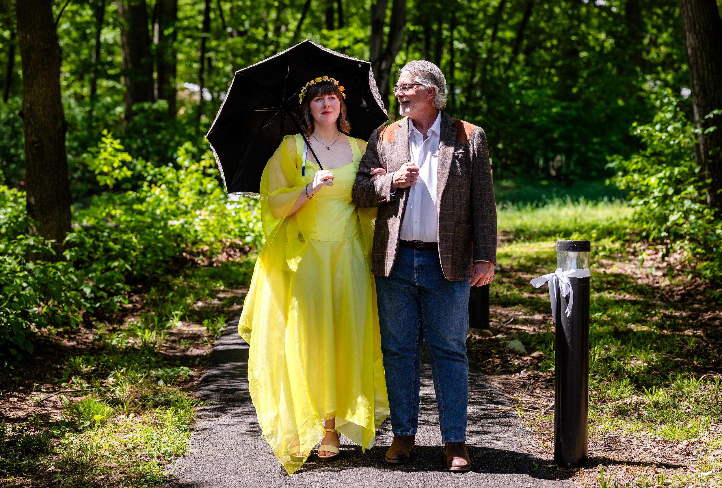 Bride in yellow gown walks with older man holding black umbrella on a wooded path.