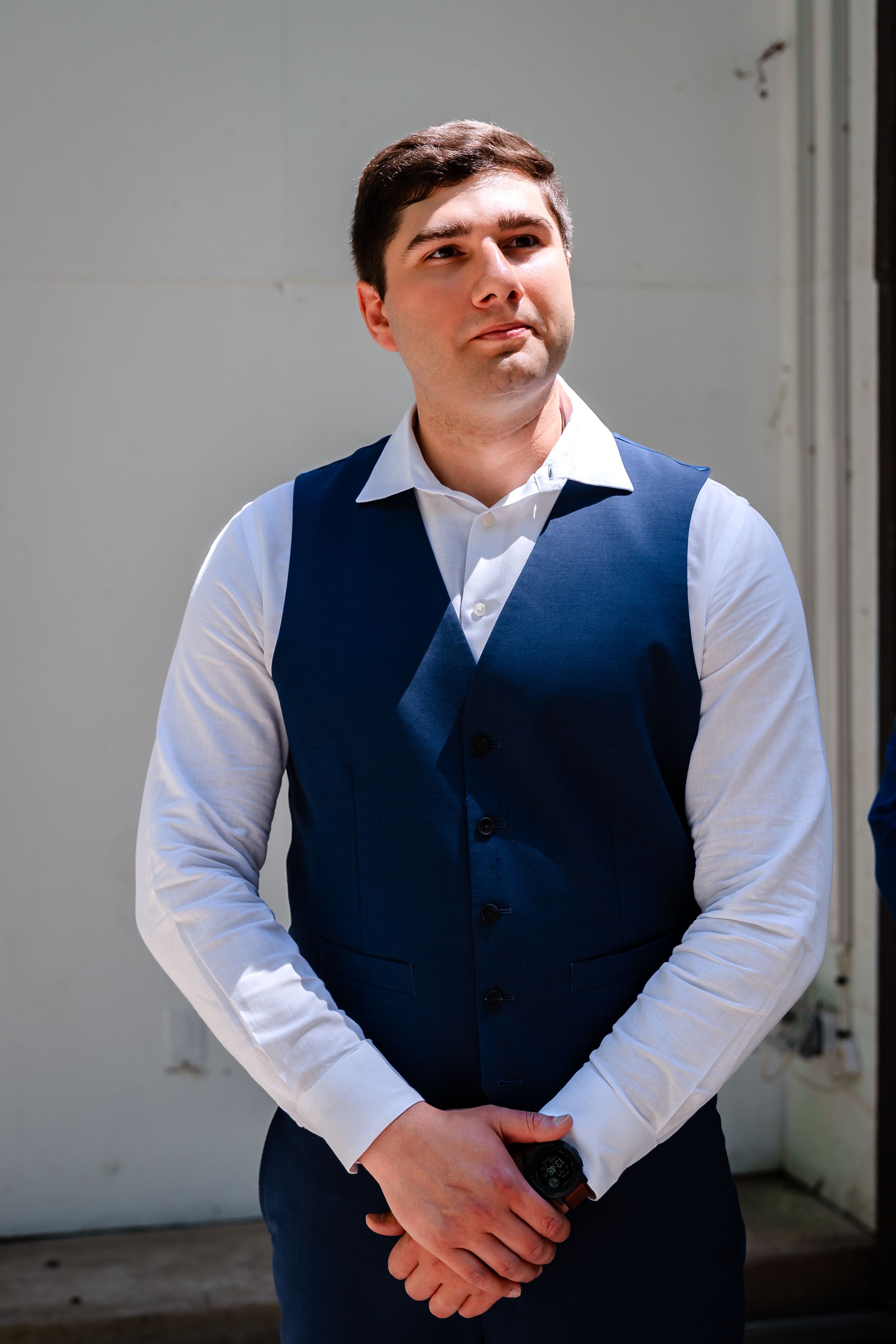 A young man in a navy blue vest and white dress shirt stands confidently with hands clasped at a wedding.