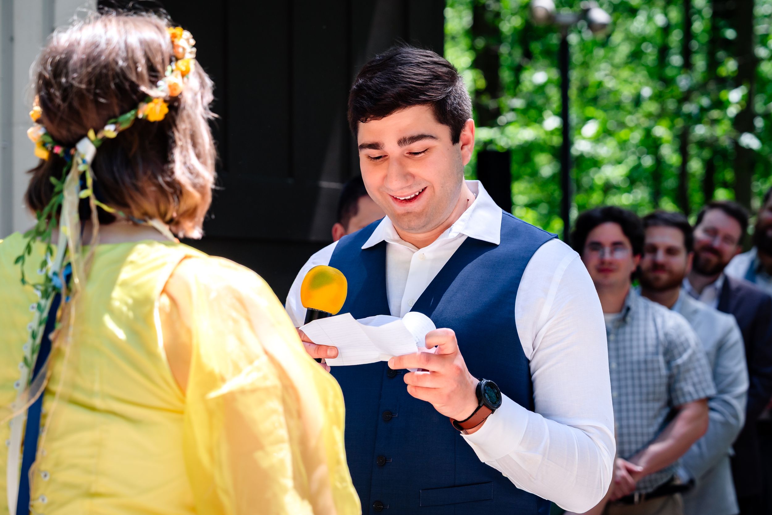 Groom in navy vest reading vows during outdoor wedding ceremony.