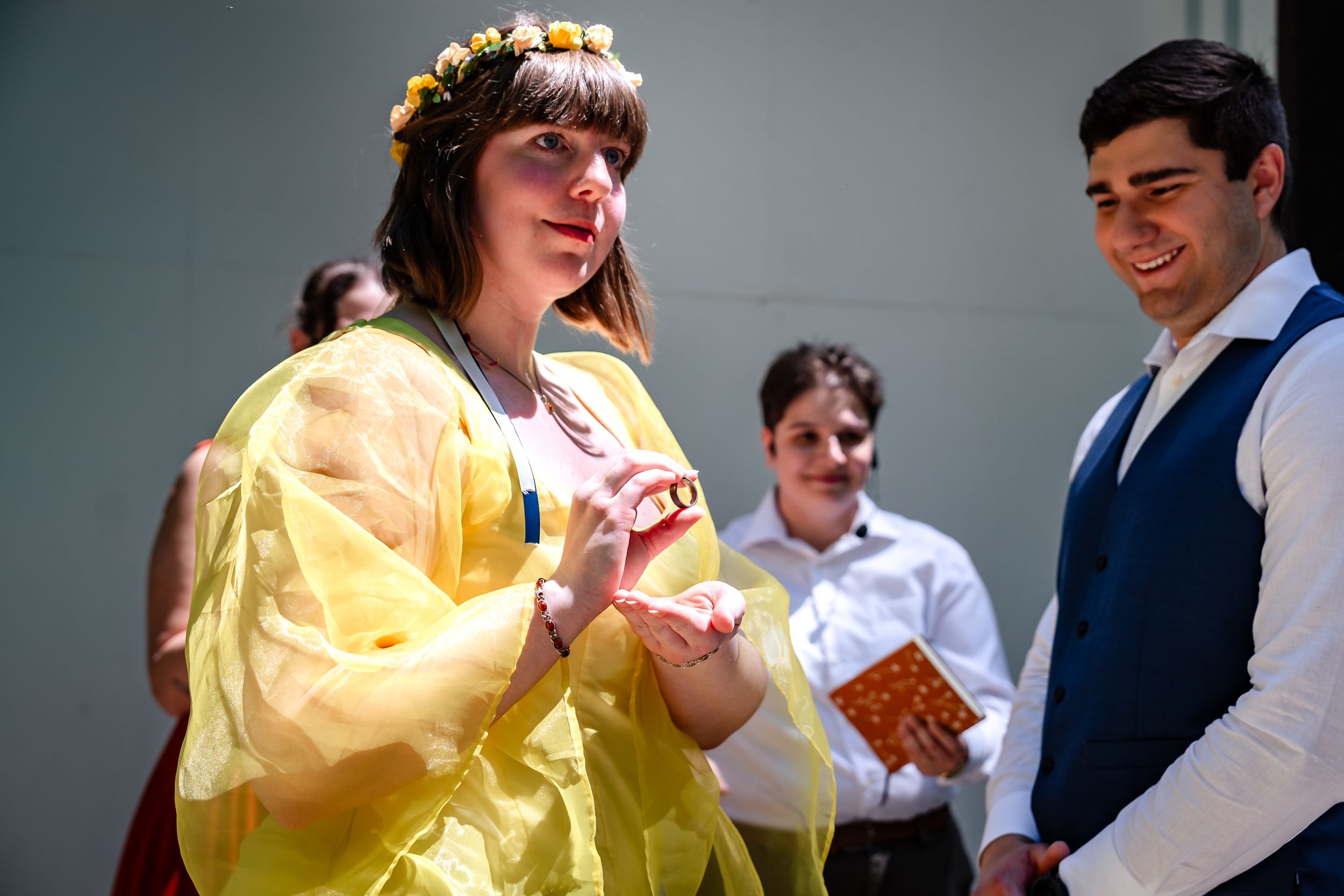Bride in yellow dress with floral crown exchanging rings at outdoor wedding.