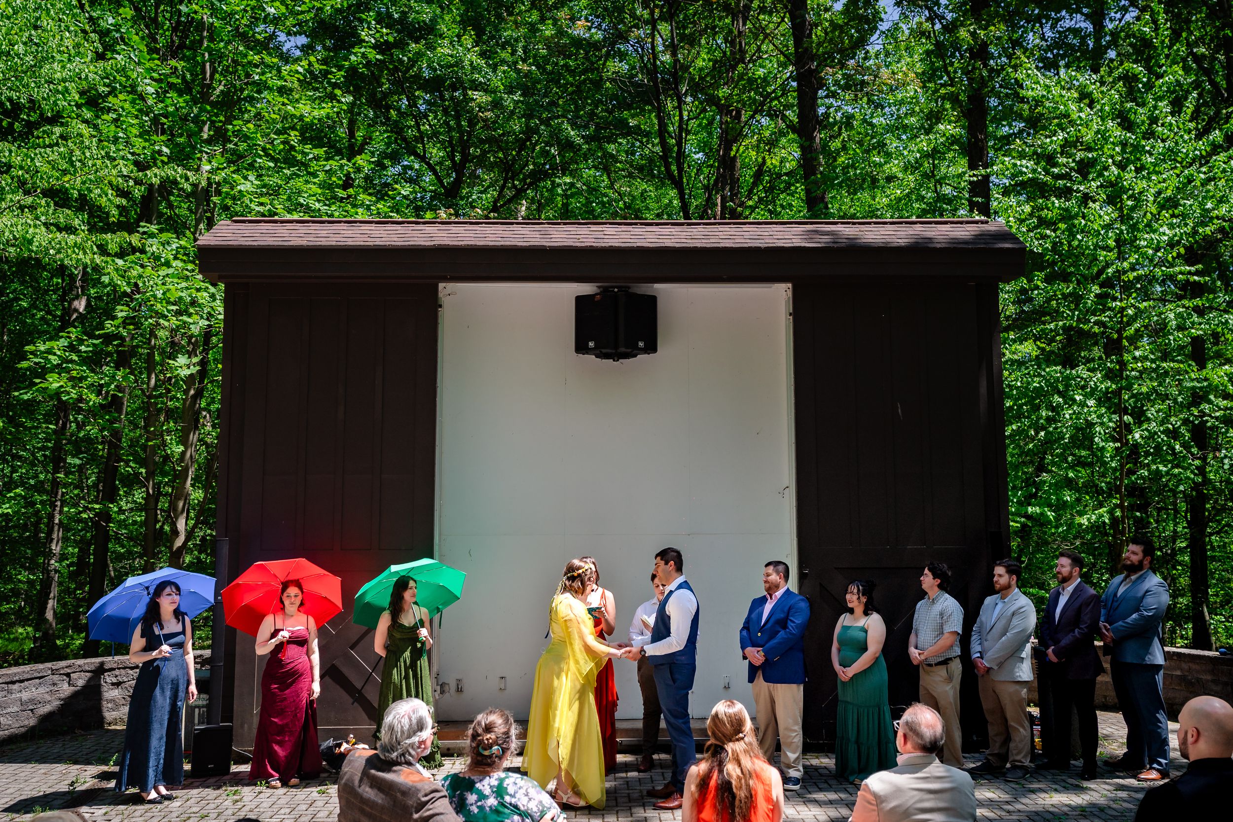 Wedding ceremony outdoors in lush green forest with colorful umbrellas and guests gathered around couple.