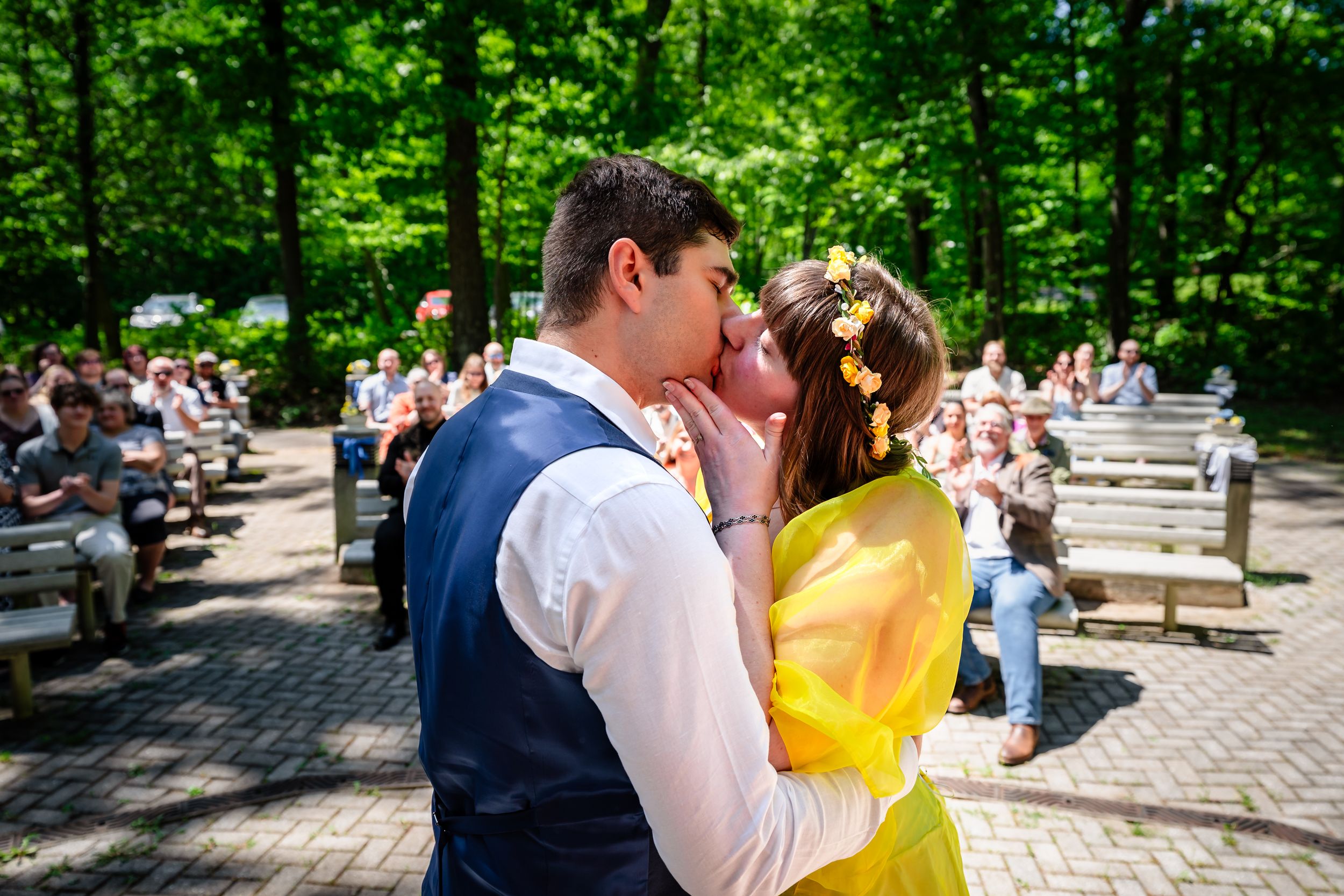 Newlyweds sharing first kiss at outdoor woodland wedding ceremony with guests seated.