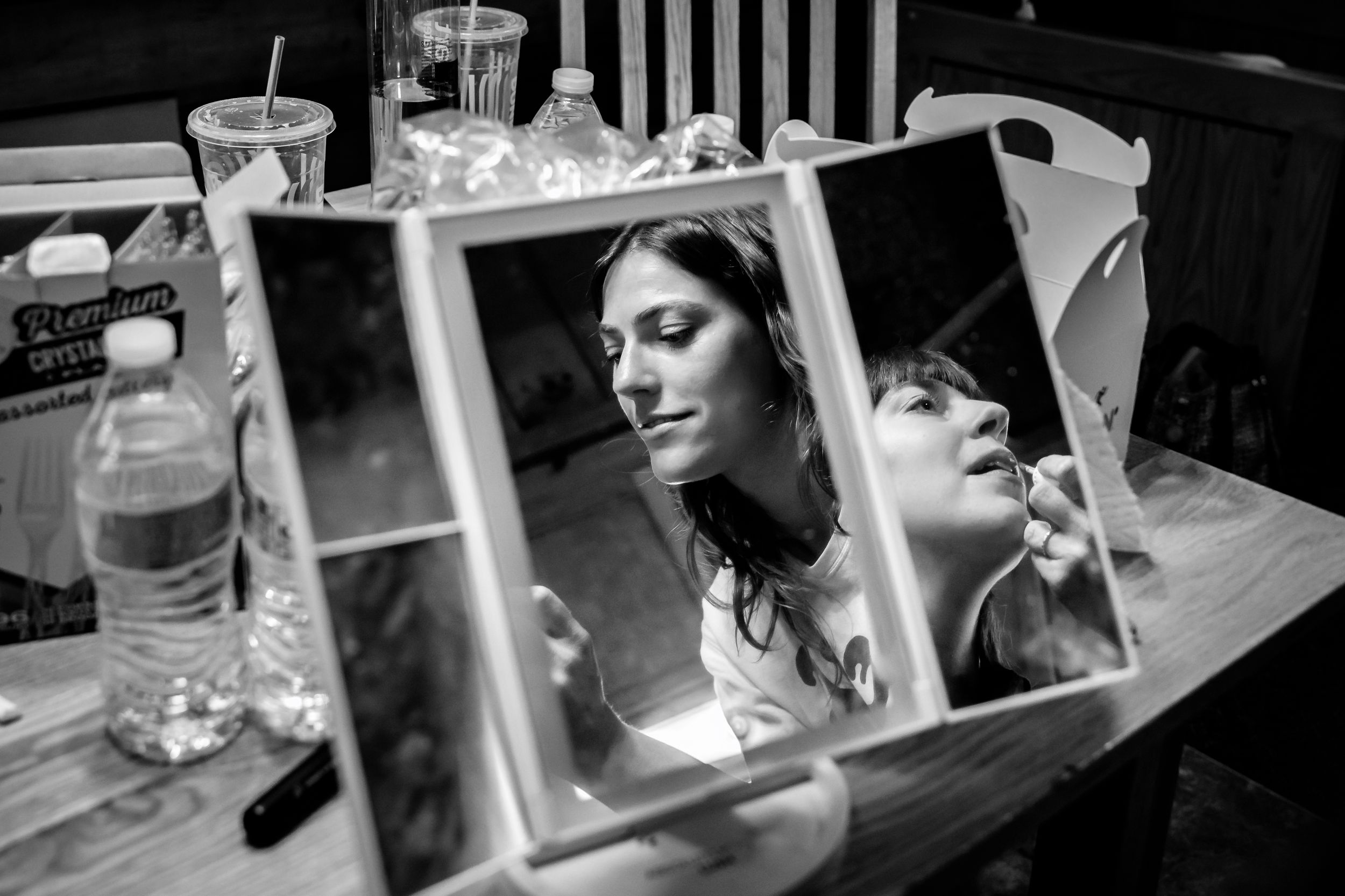 Black and white photo of a woman applying makeup reflected in a trifold vanity mirror on a table.