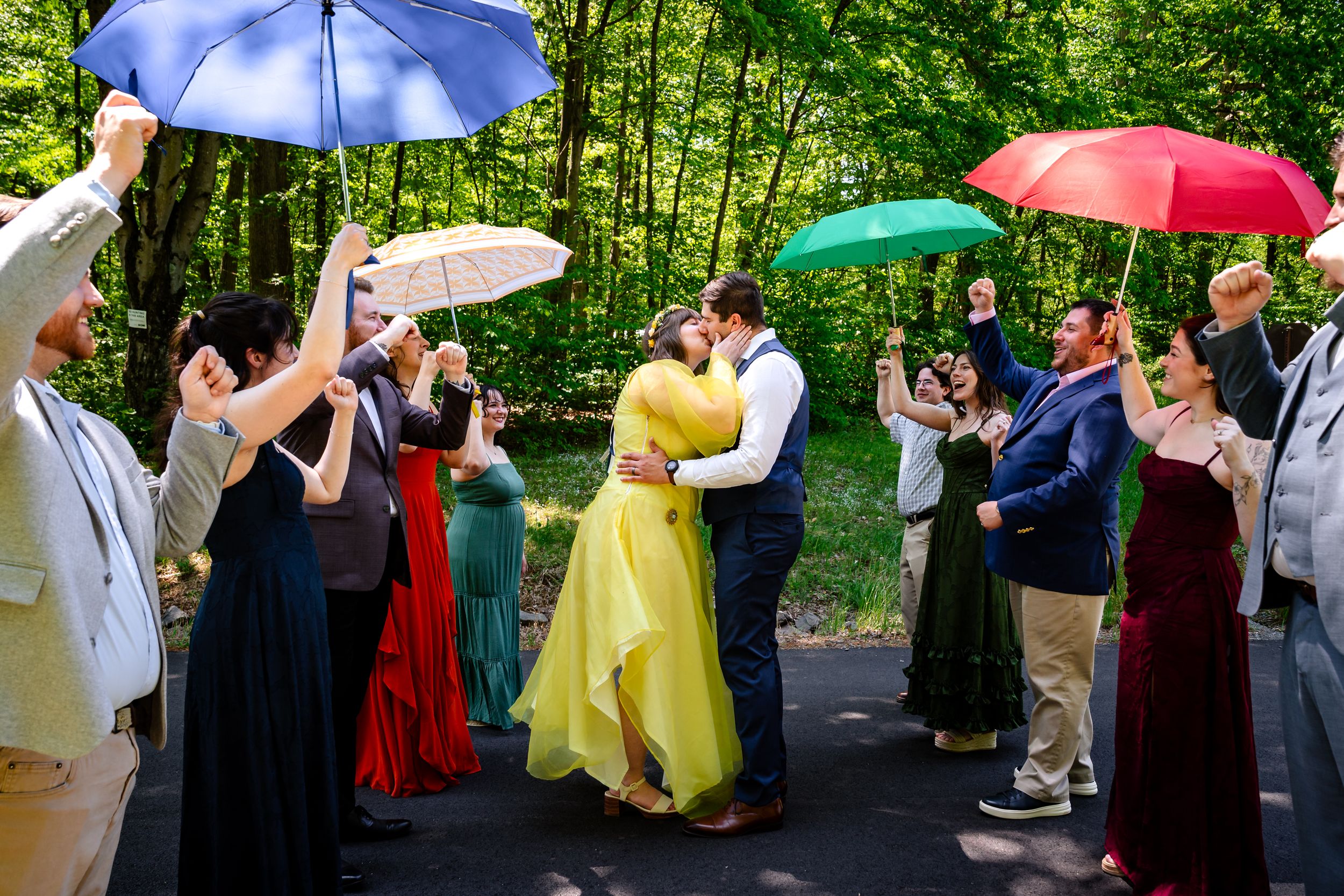 Wedding couple shares first kiss under colorful umbrellas held by guests in lush green forest.