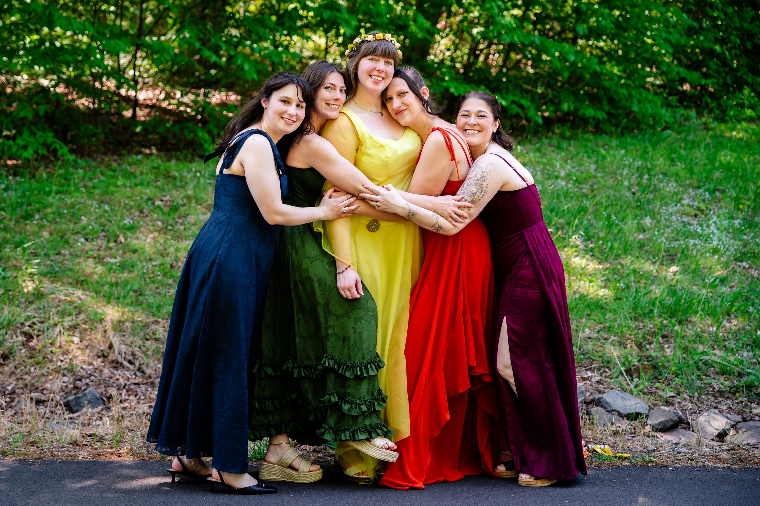 Bride in yellow gown embraces four bridesmaids in rainbow-colored dresses outdoors in nature.