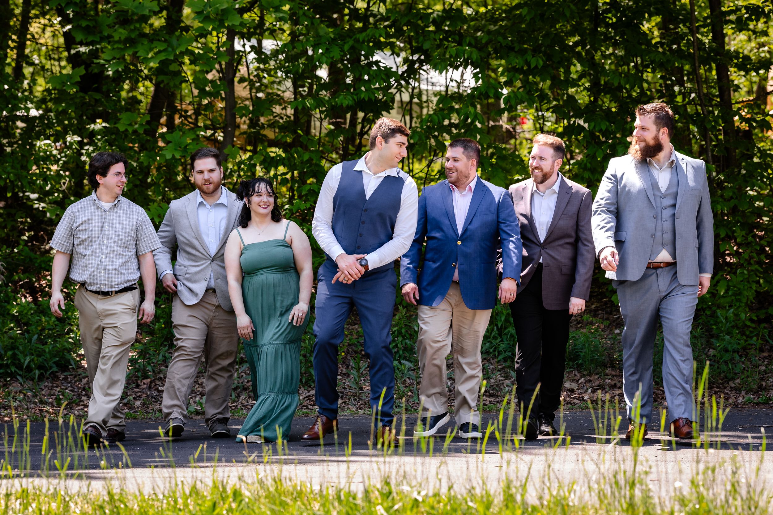 Wedding party walks together outdoors among lush green trees on a sunny day.