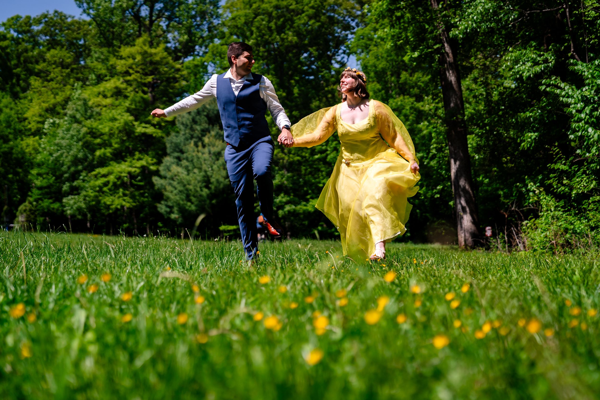 Bride in yellow gown and groom in blue suit run joyfully through sunny green meadow with wildflowers.