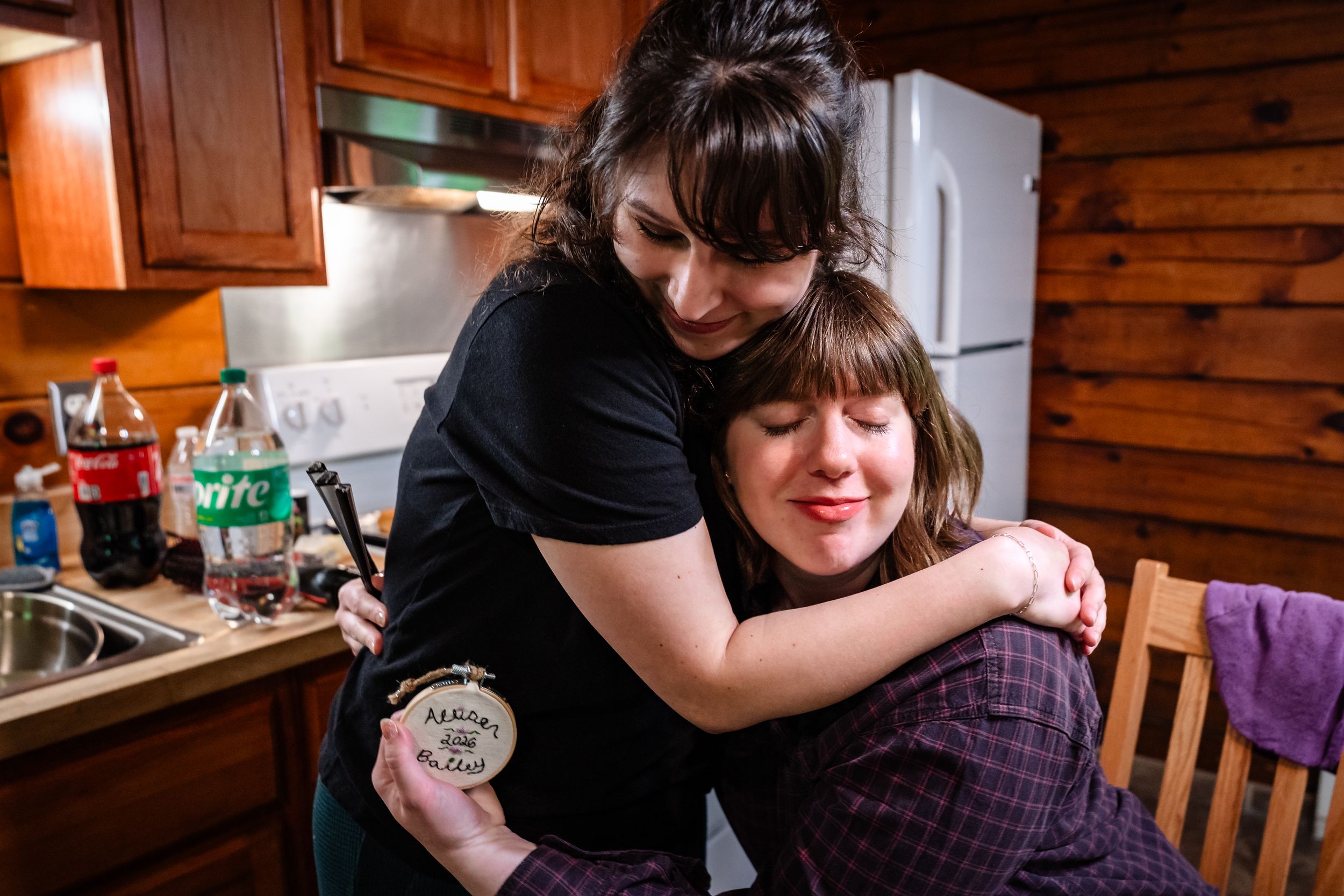 Two women share a warm embrace in a cozy kitchen, one holding a decorated jar.