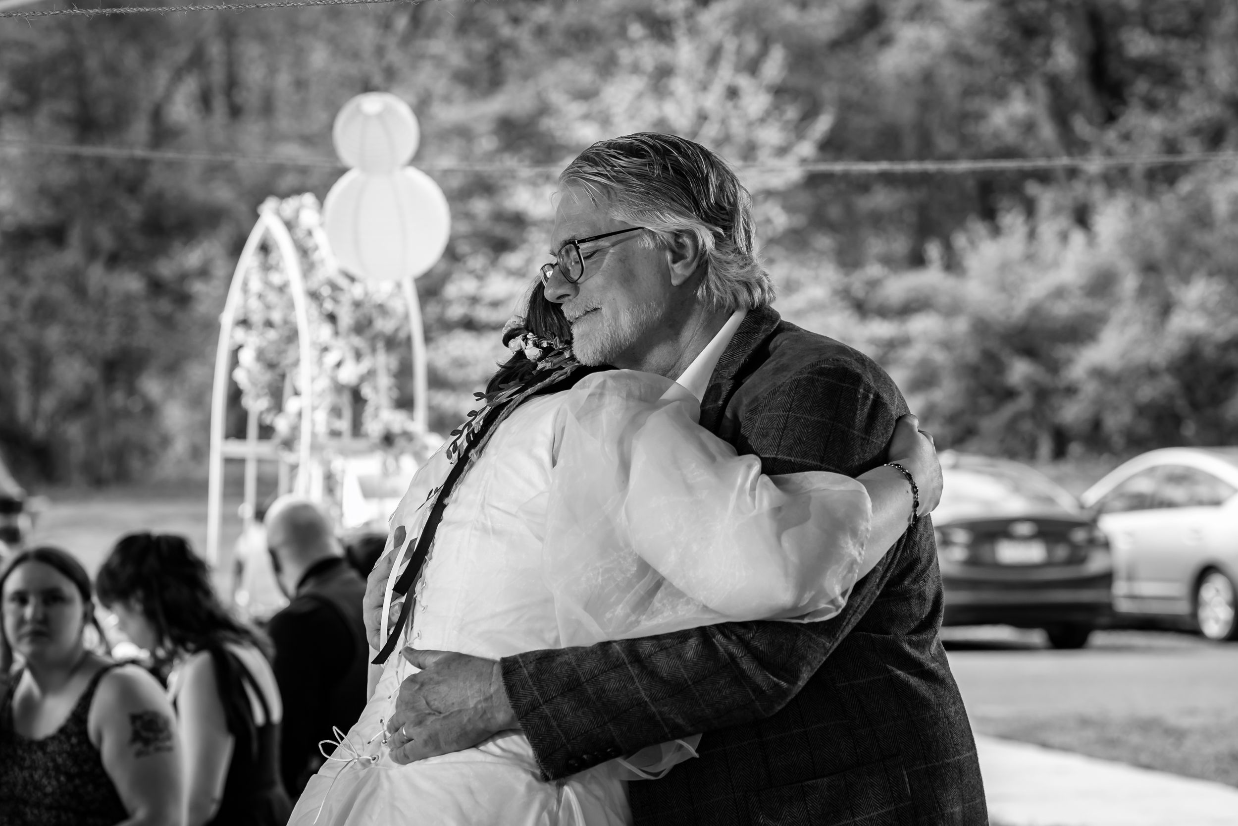Emotional father hugs bride in white wedding dress at outdoor ceremony in black and white photo.