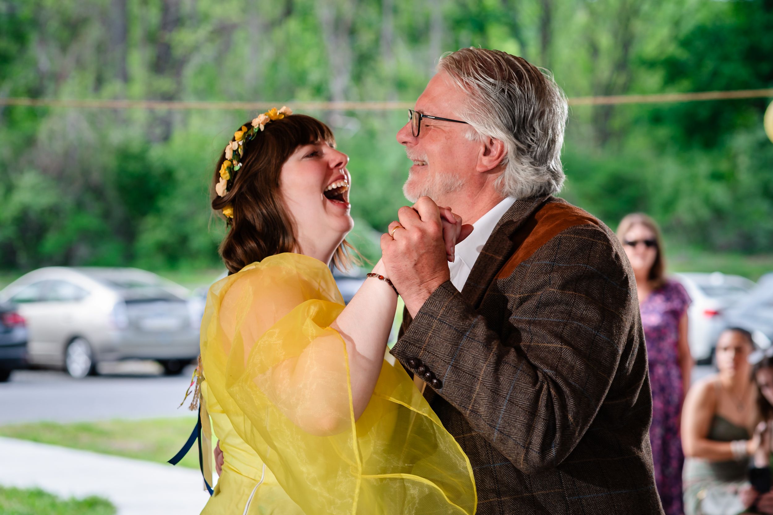 Bride in yellow dress laughs joyfully while dancing with older man in brown suit at outdoor wedding.