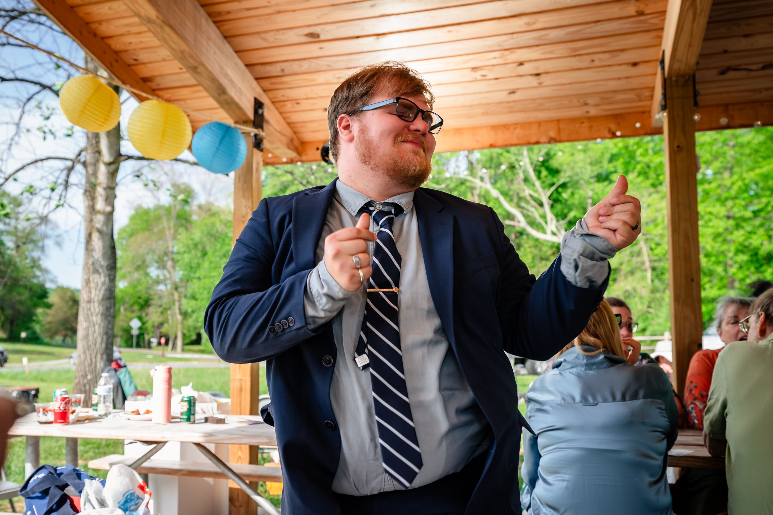 Man in navy blazer and striped tie dancing joyfully under decorated pavilion.