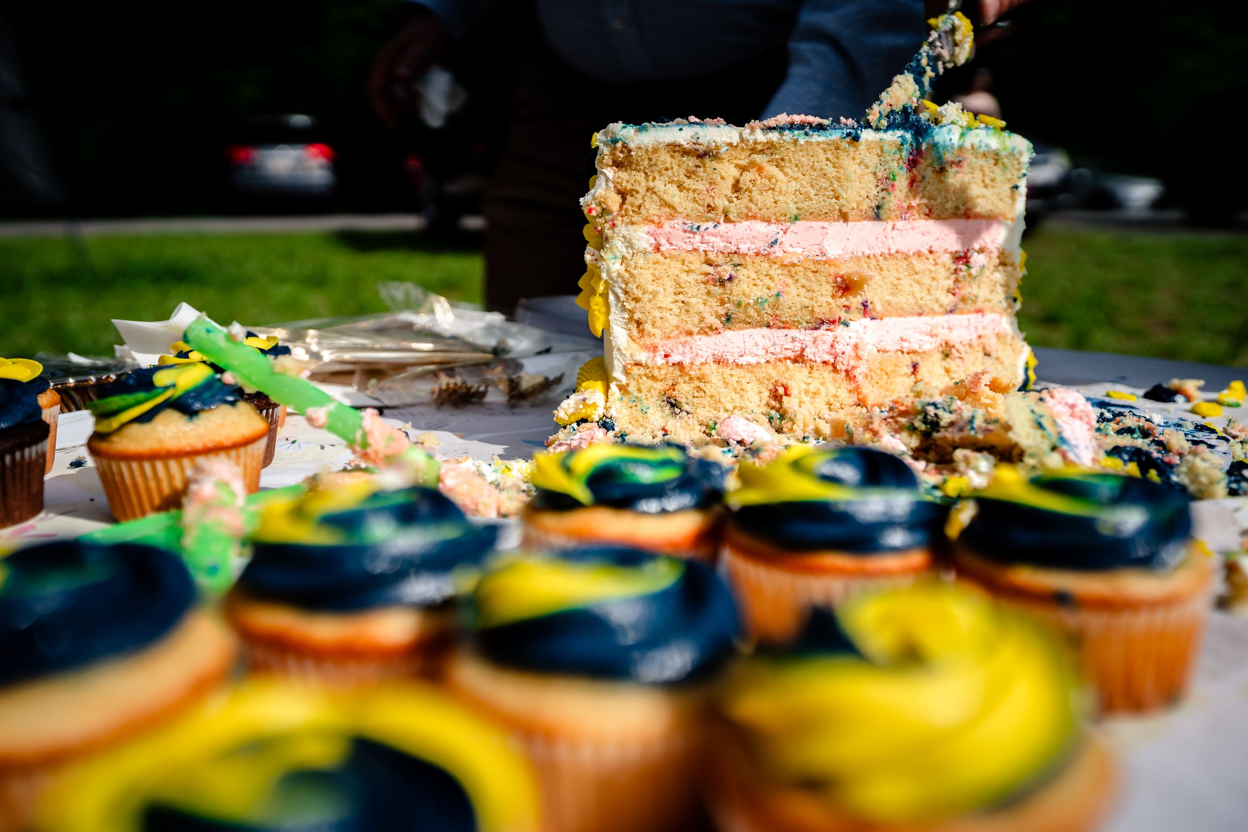 Colorful wedding celebration cake with yellow layers and cupcakes with teal and yellow frosting displayed on an outdoor table.