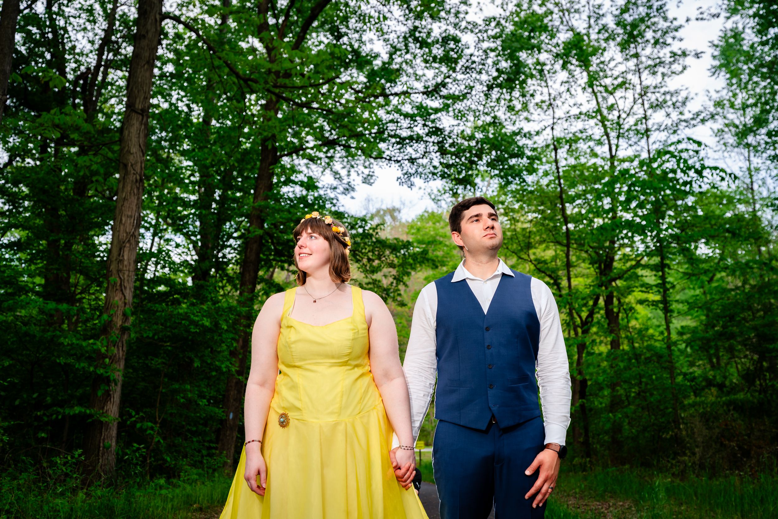 A couple in formal attire holds hands outdoors among lush green trees.