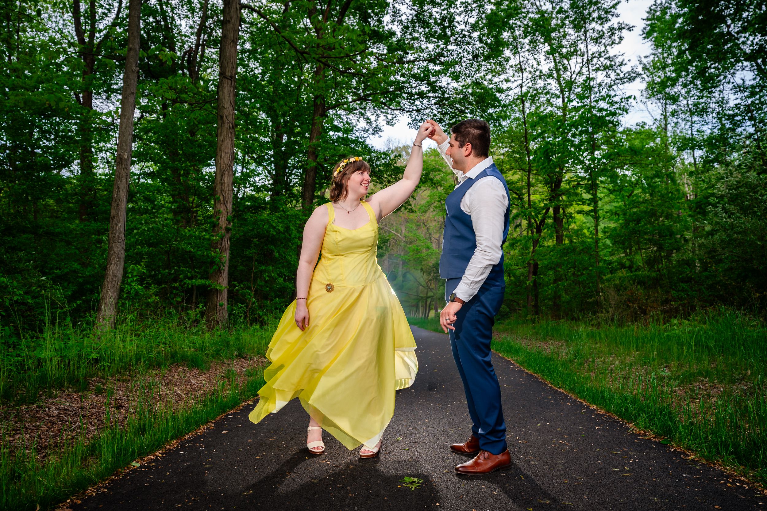 Bride and groom dance joyfully on a forest path, her yellow gown spinning in the air.