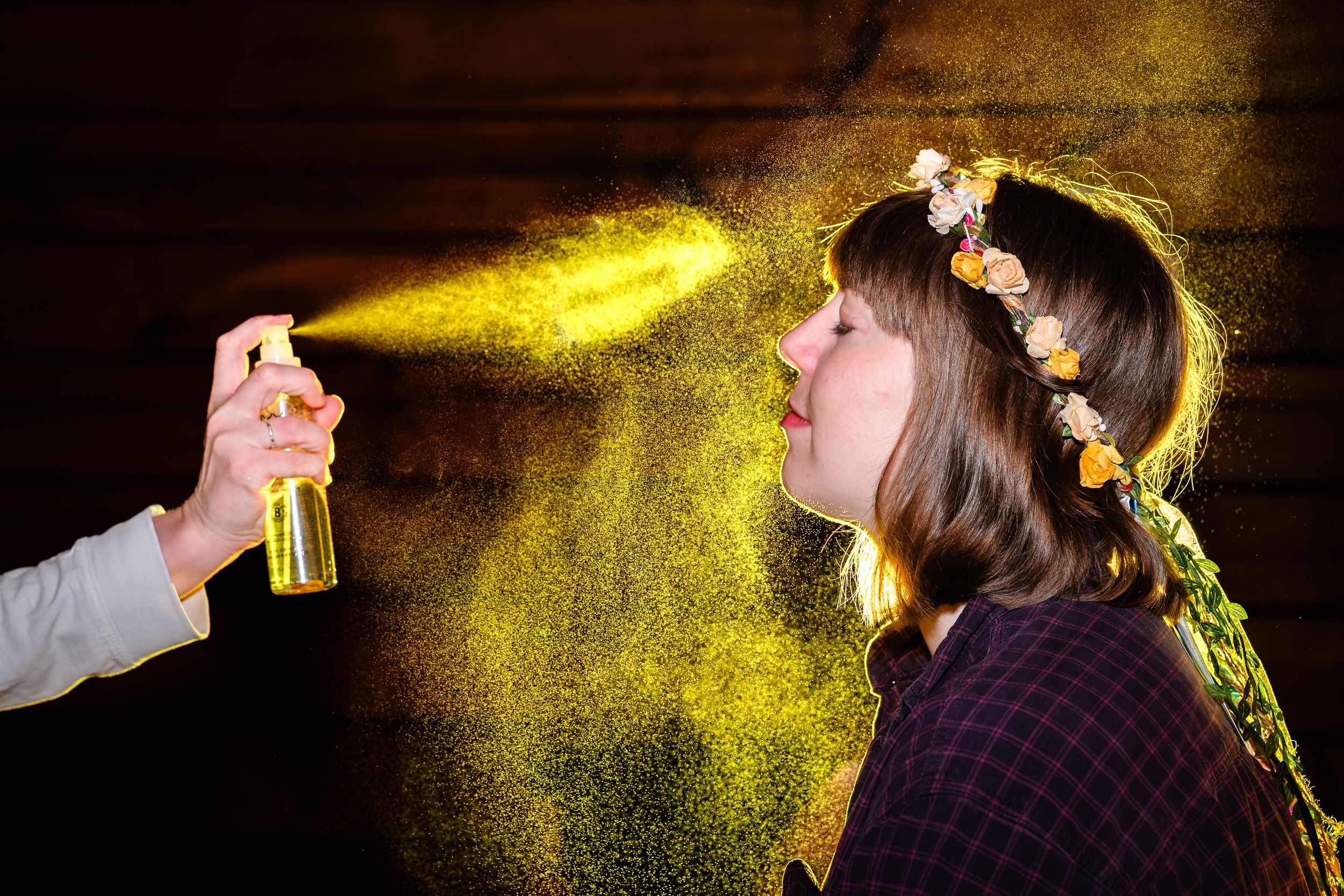 Woman with floral crown inhales golden glitter spray mist against dark background in artistic portrait photo.