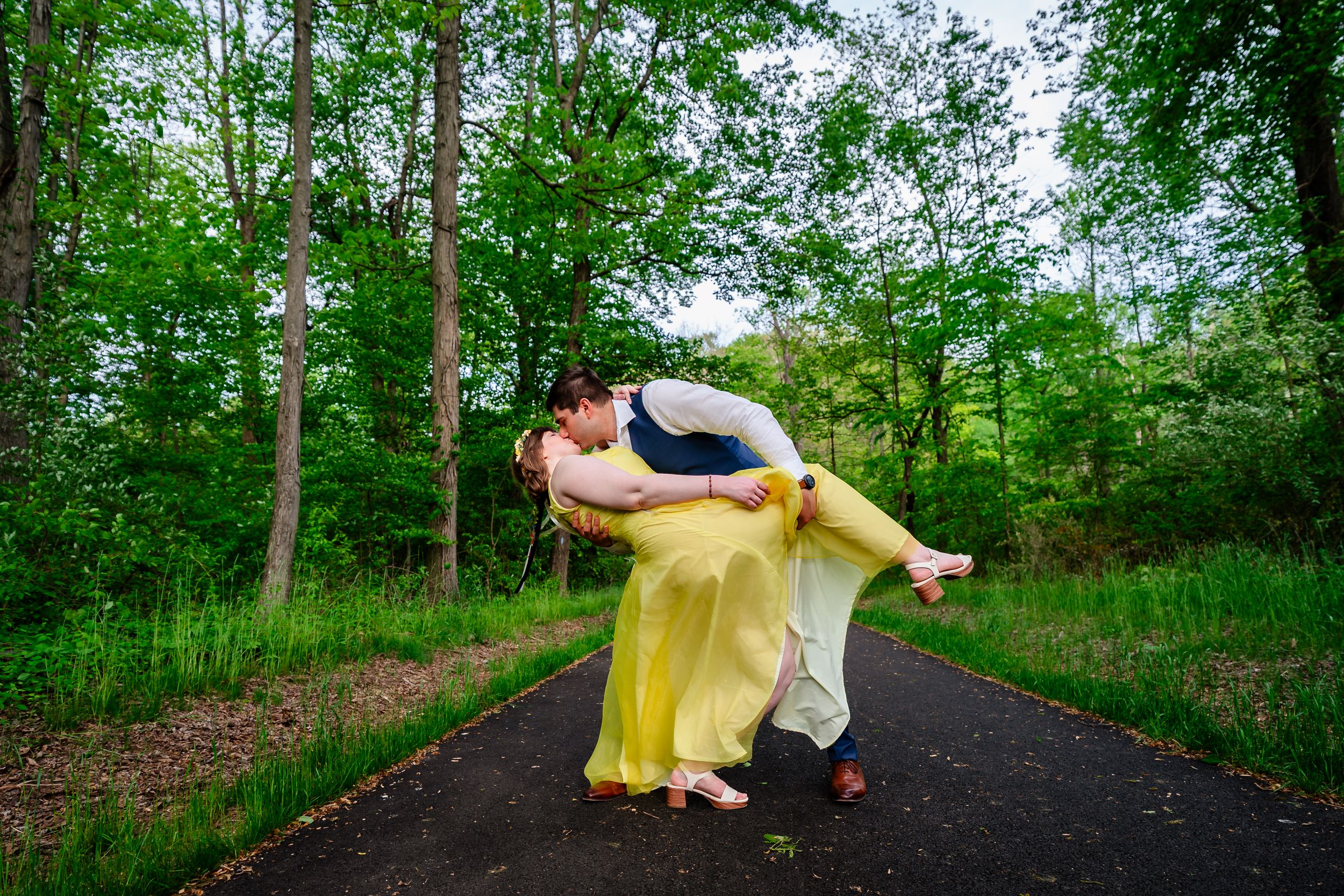 Groom dips bride in a flowing yellow dress for a romantic kiss on a tree-lined path.