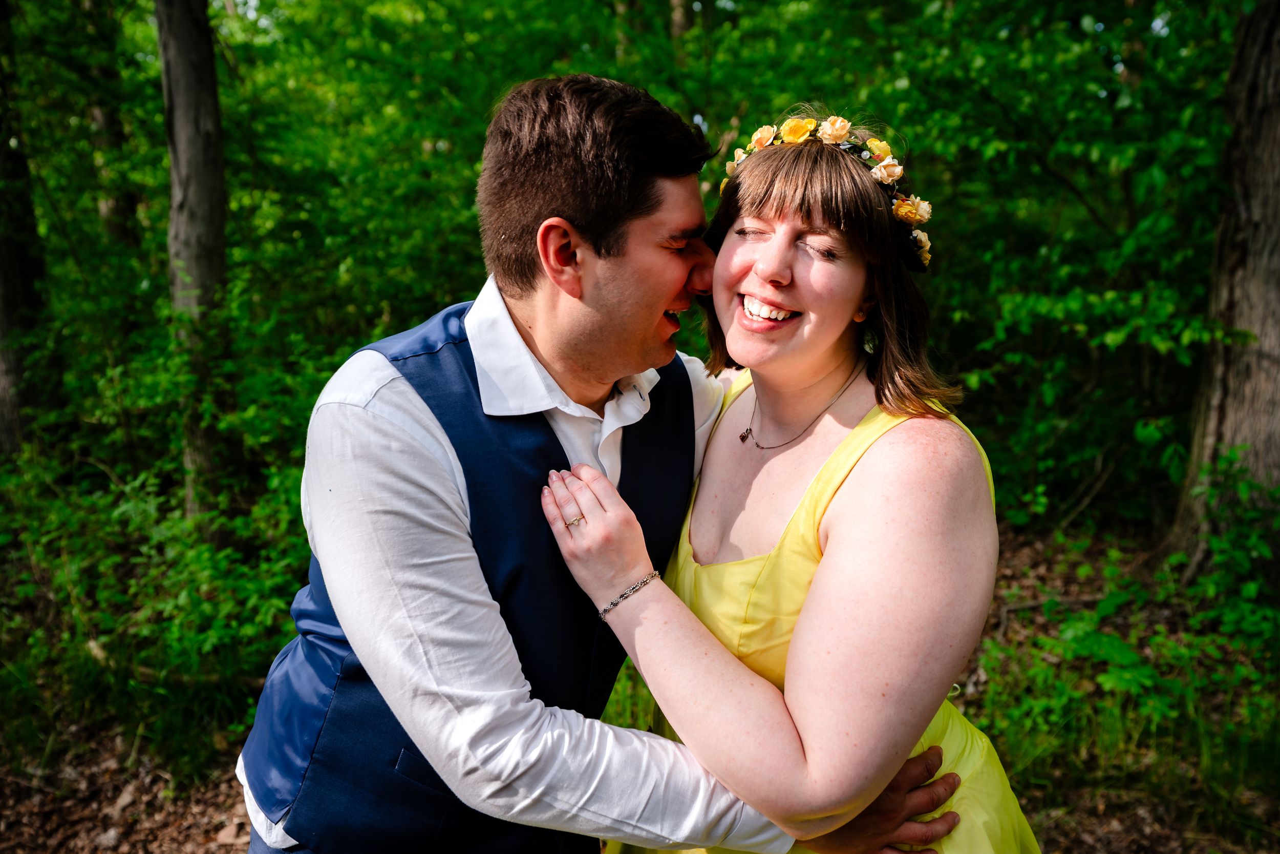 Laughing bride in yellow dress and floral crown embracing groom in forest setting.