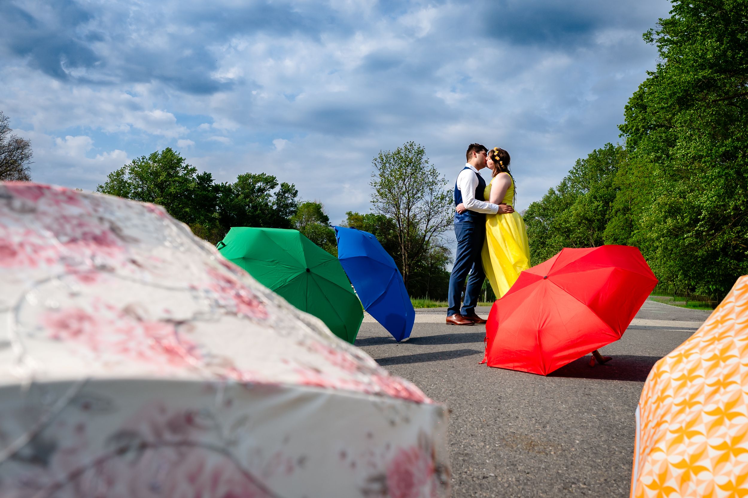 Bride and groom kiss surrounded by colorful open umbrellas on a scenic road under blue skies.