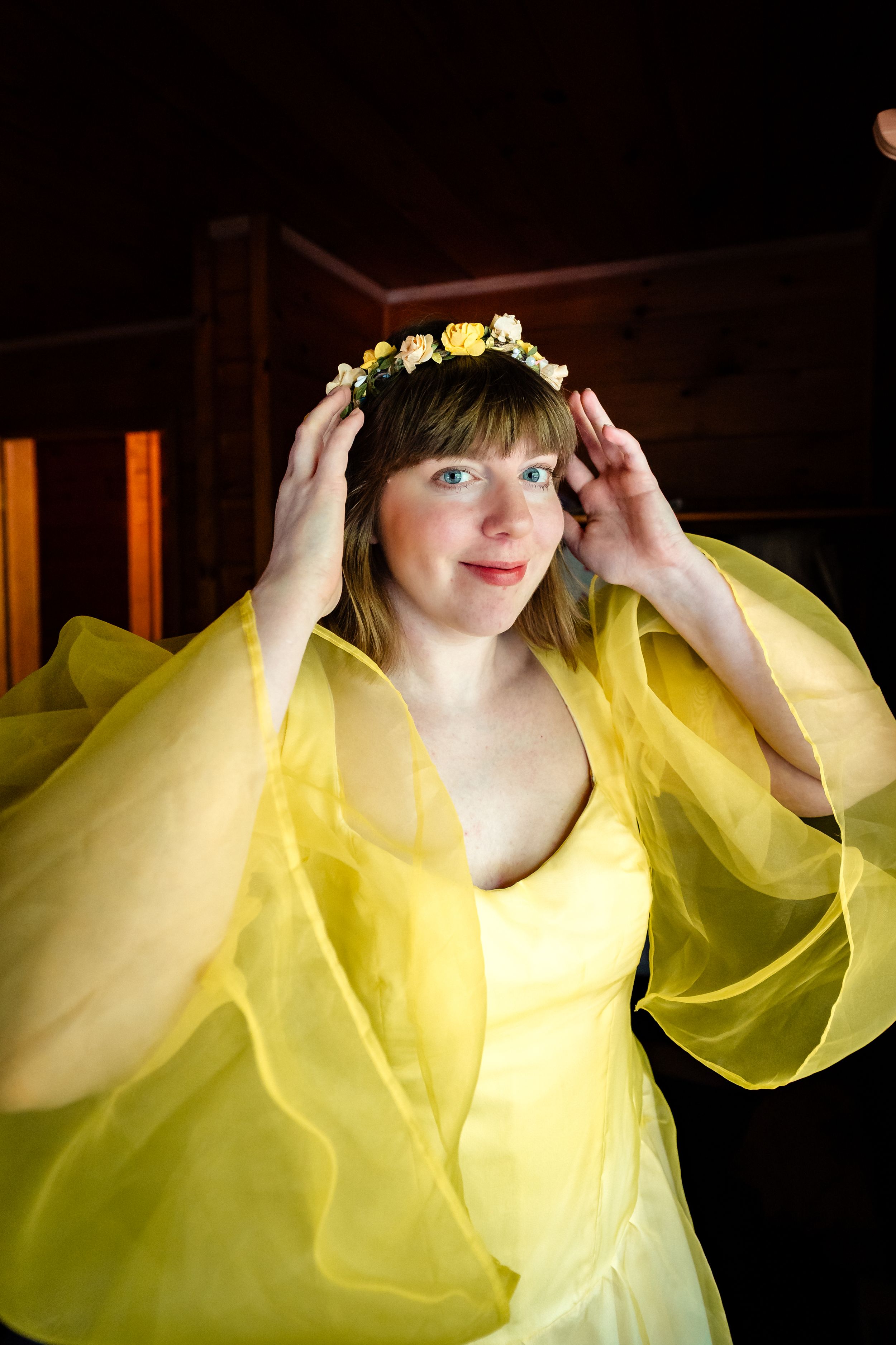 Woman in yellow gown and floral crown poses elegantly against dark background.