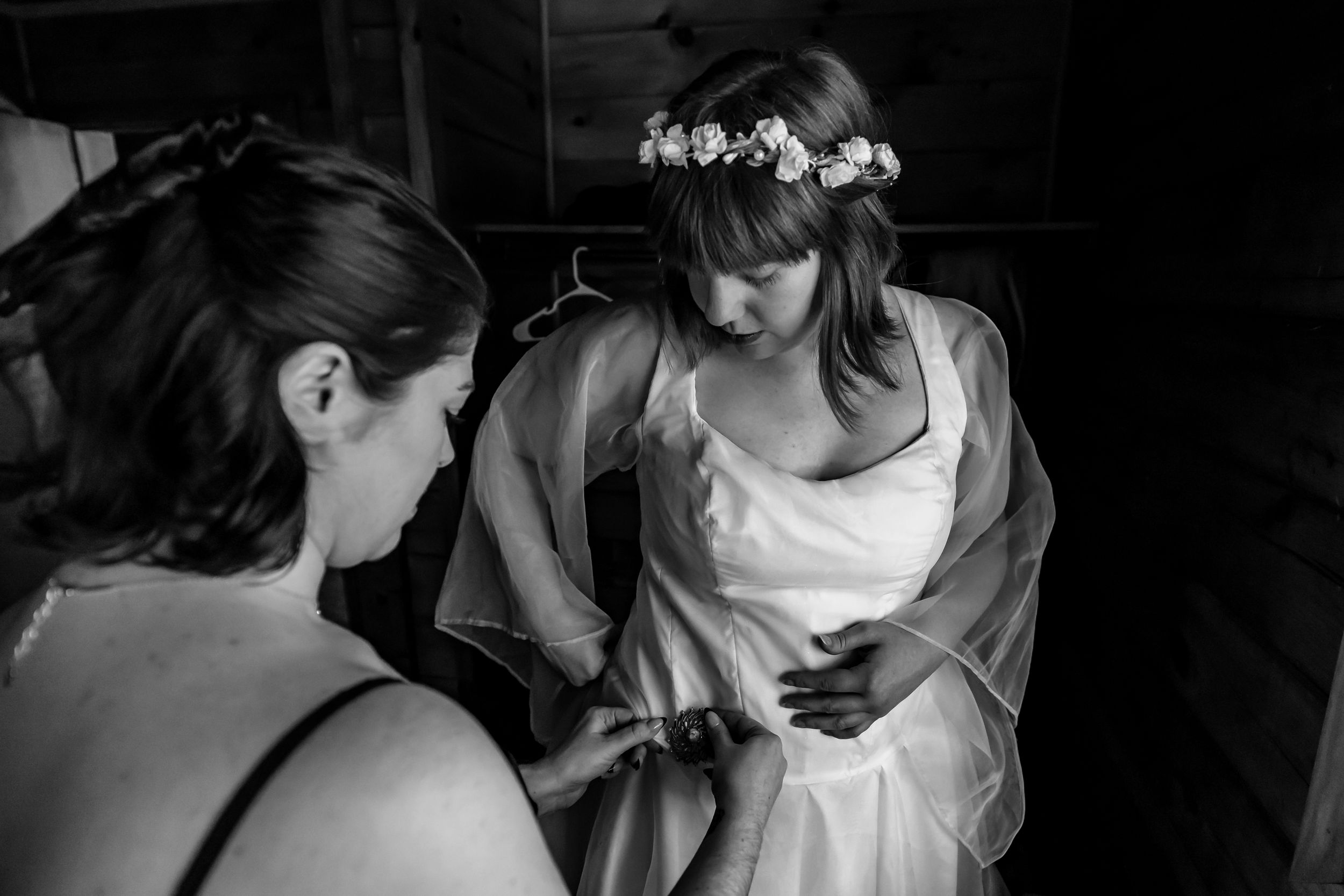 Bride in white gown and floral crown gets help from bridesmaid in black-and-white wedding preparation photo.