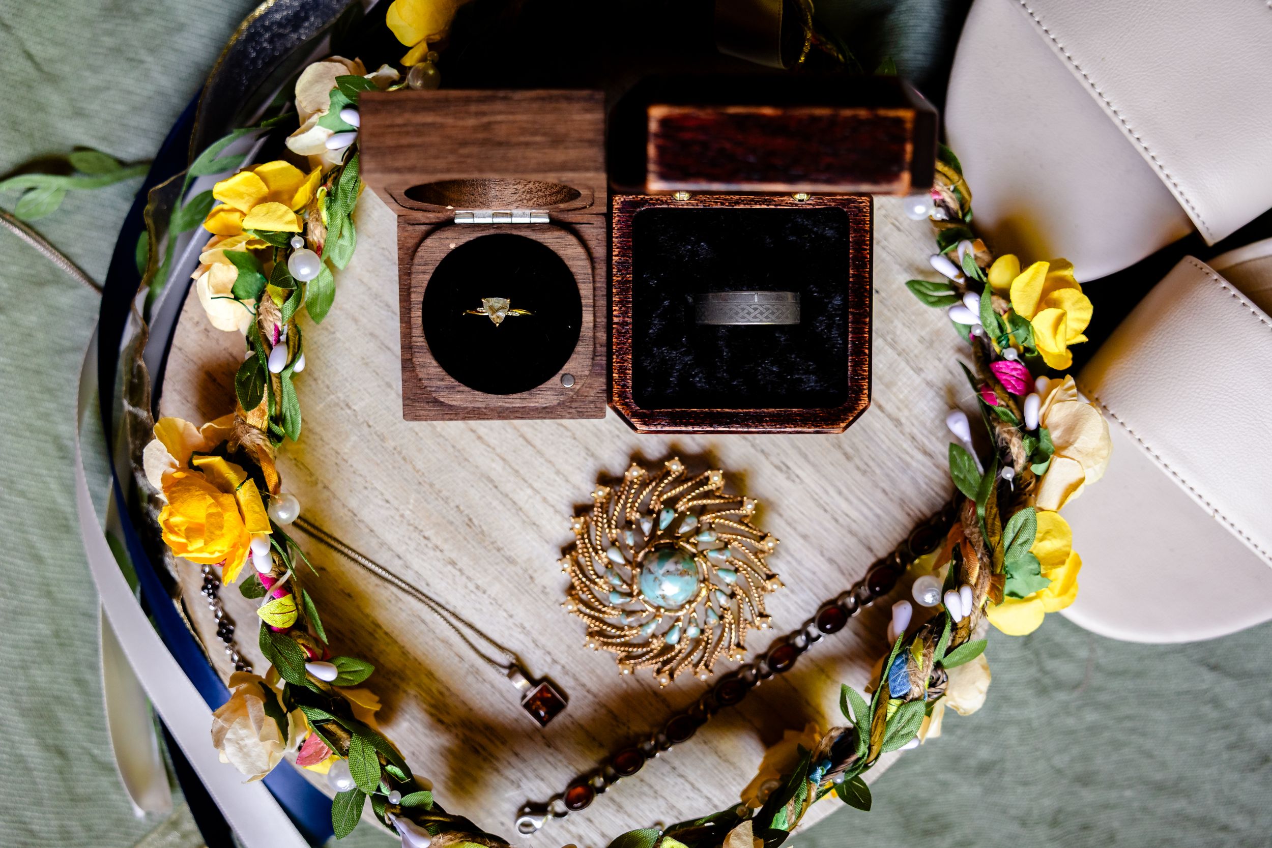 Wedding rings displayed in wooden box with floral crown and decorative brooch on white textured surface.