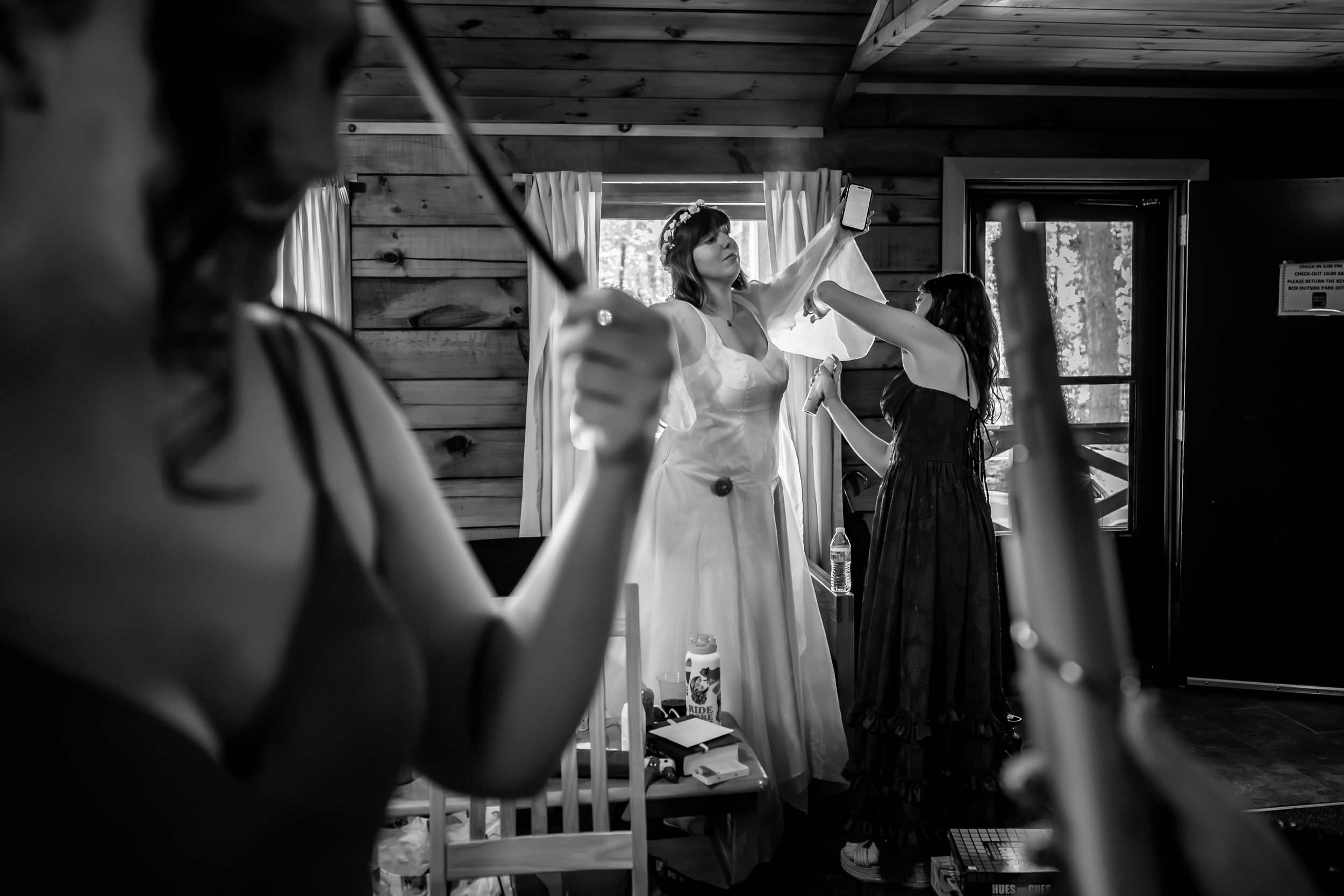 Black and white photo of bridesmaids helping bride into her wedding dress inside a rustic log cabin.
