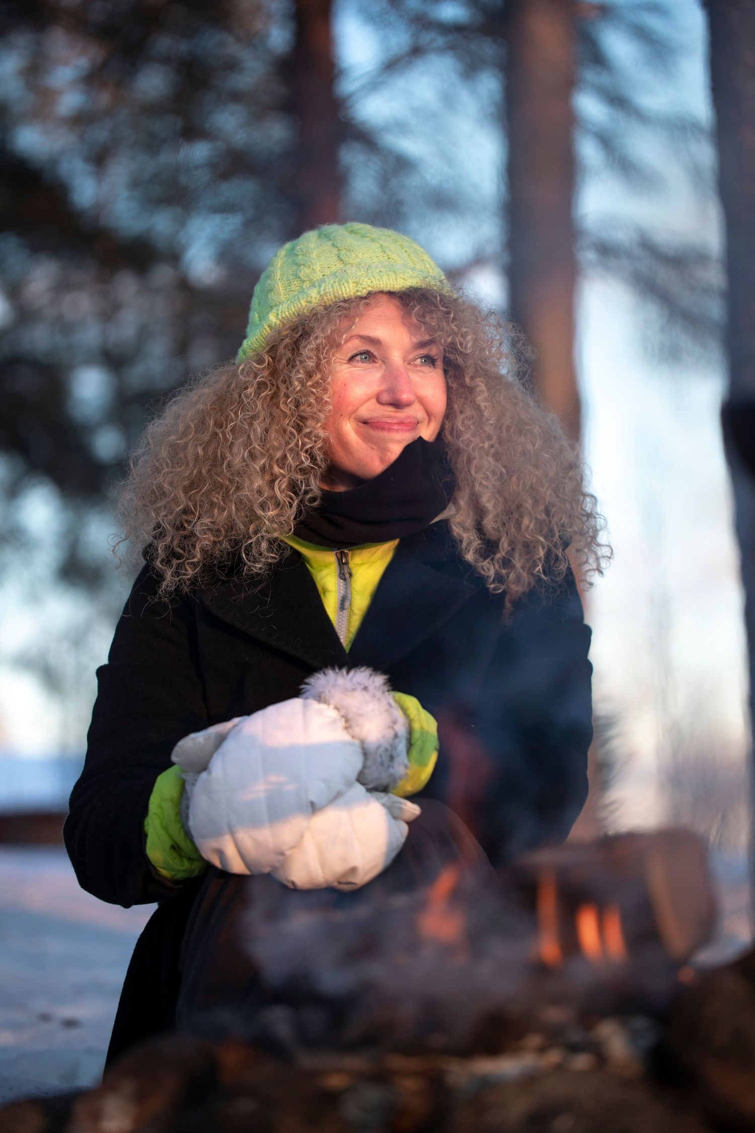 Curly-haired woman in green hat and white mittens smiling while warming hands by a winter campfire.