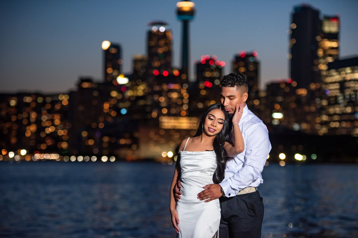 Couple embracing by the waterfront with a glowing city skyline at night.