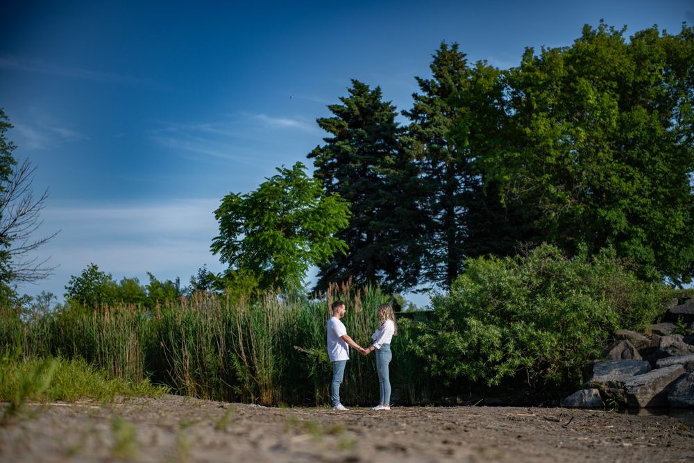 Couple holding hands on a sandy path with tall green trees under a blue sky.