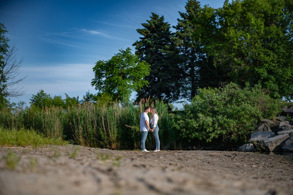 Couple standing close together on a sandy path surrounded by tall grass and trees.