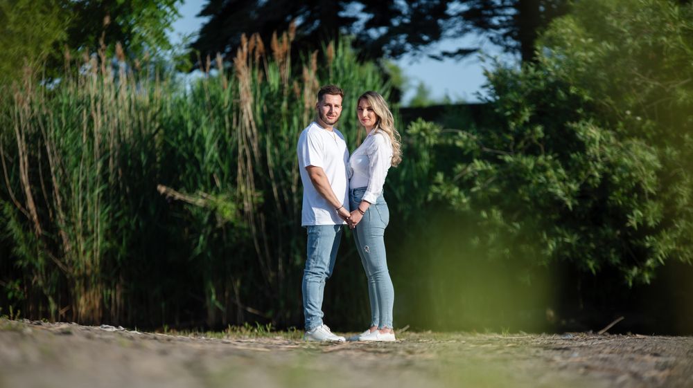 Smiling couple holding hands on a sandy path with tall green reeds behind them.
