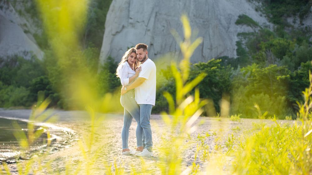 Couple holding each other closely in a golden sunlit field near a rocky cliff and stream.