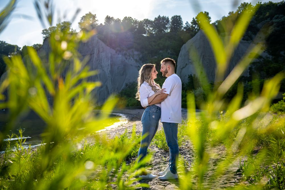 Couple embracing outdoors near rocky cliffs with sunlight streaming through green foliage.