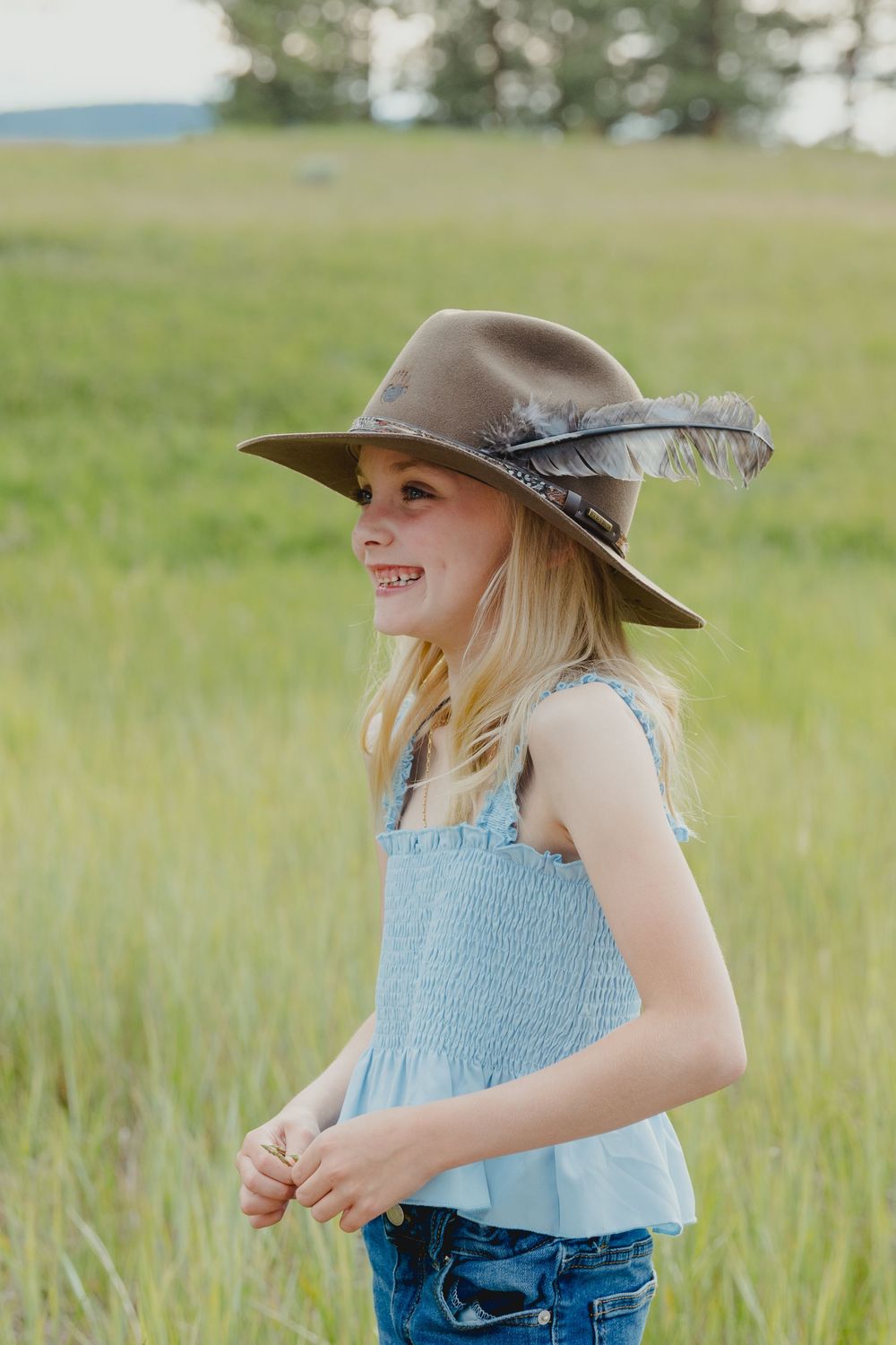 A smiling blonde girl wearing a wide-brim hat with feathers and a blue top stands in a lush green field.