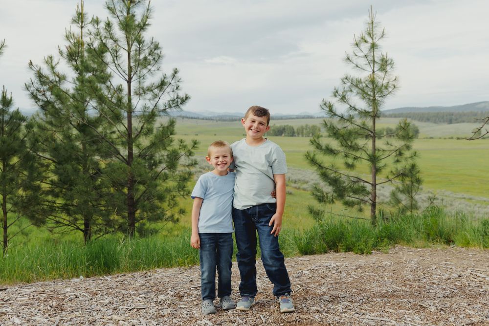 Two young boys smiling and posing together outdoors near pine trees with green meadows and mountains in the background.