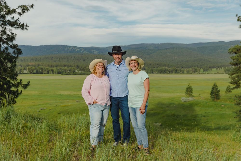 Three people in cowboy hats smiling in a lush green meadow with mountain forest backdrop.