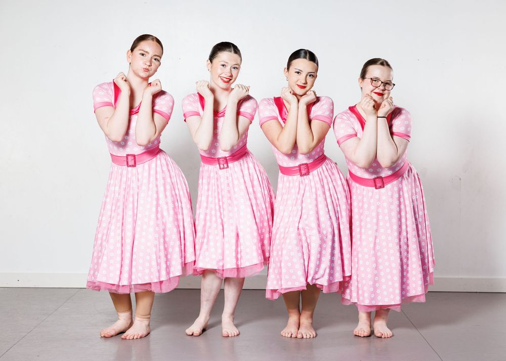 Four girls in matching pink polka dot dresses with pink belts pose together barefoot against a white wall.