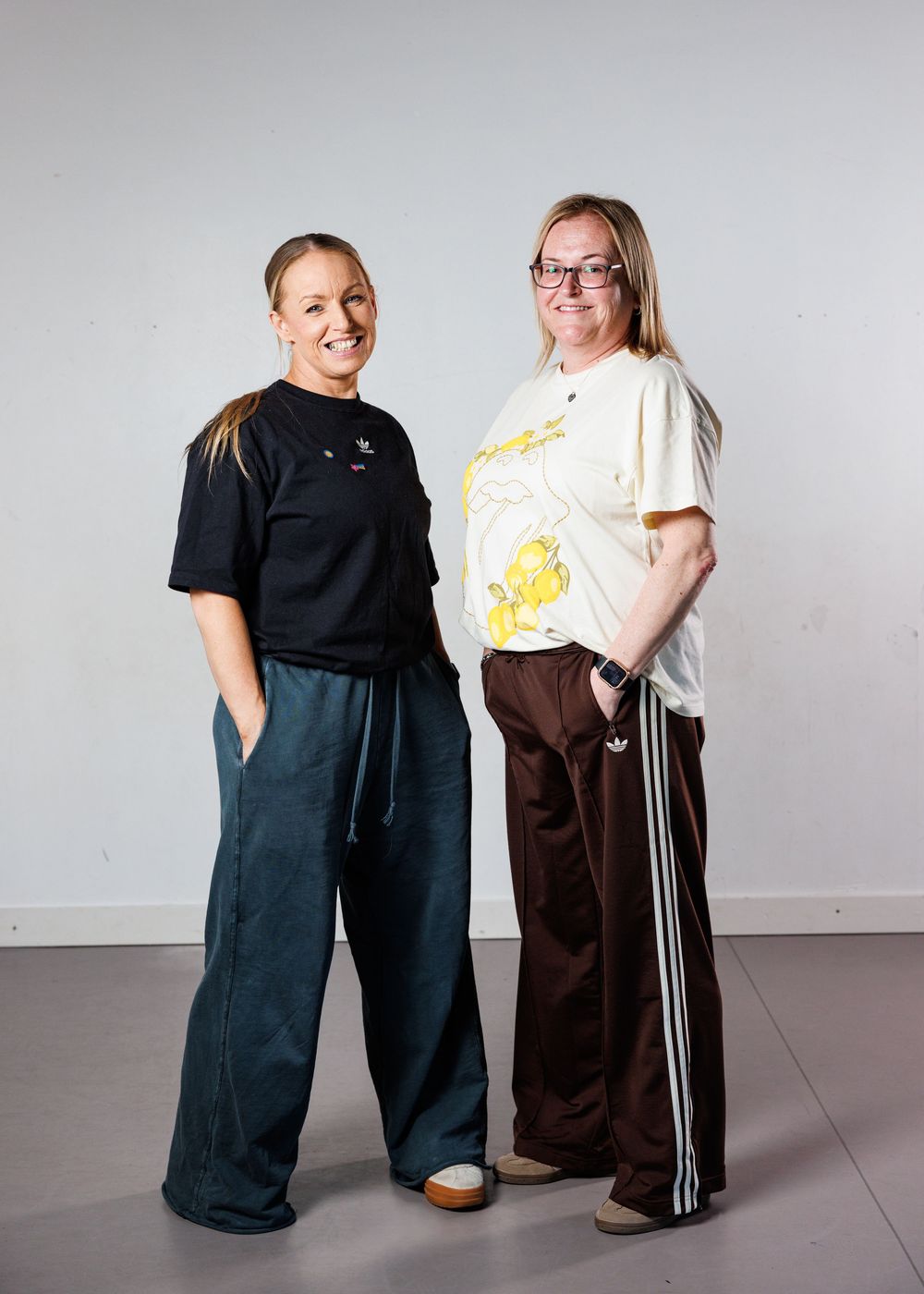 Two women in casual Adidas sportswear pose together against a light gray background.