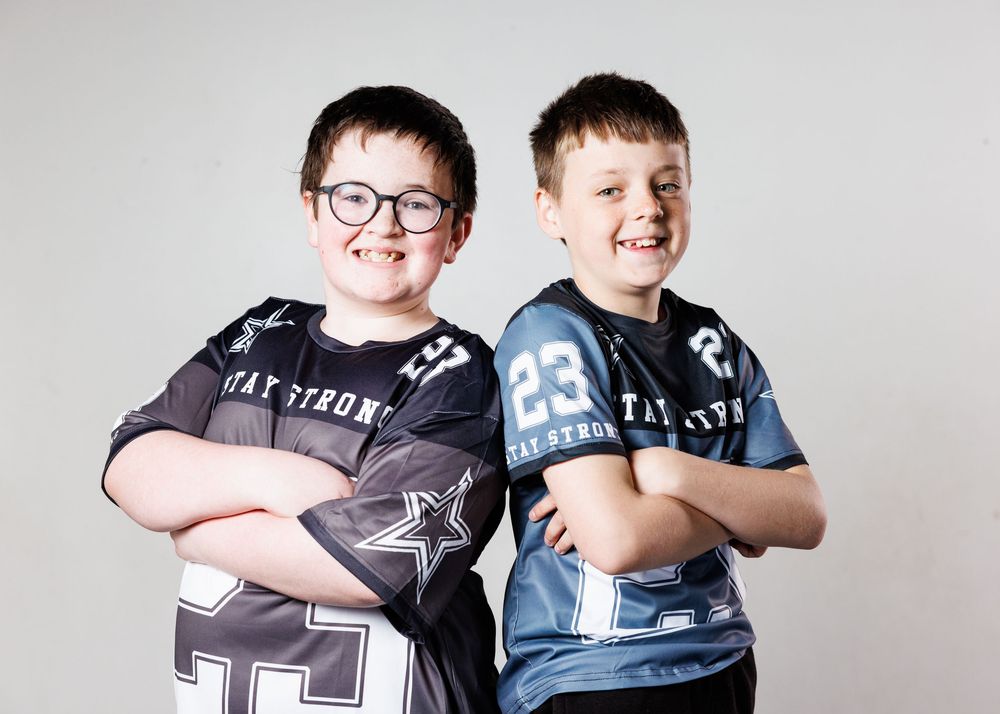 Two boys in matching Stay Strong football jerseys pose with arms crossed, smiling confidently against a gray background.