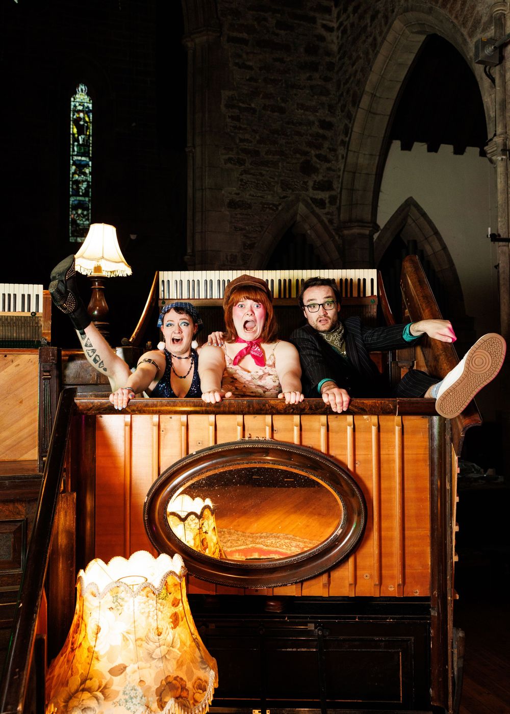 Three quirky performers pose playfully in a wooden church pulpit, with gothic arches and stained glass in the background.