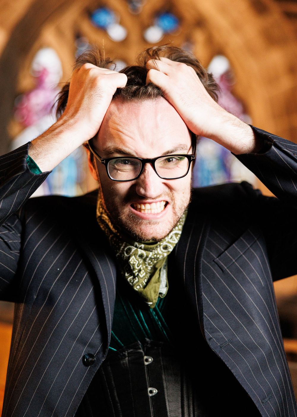 Man in pinstripe suit and glasses gripping his hair with an intense expression, standing before a stained glass window.