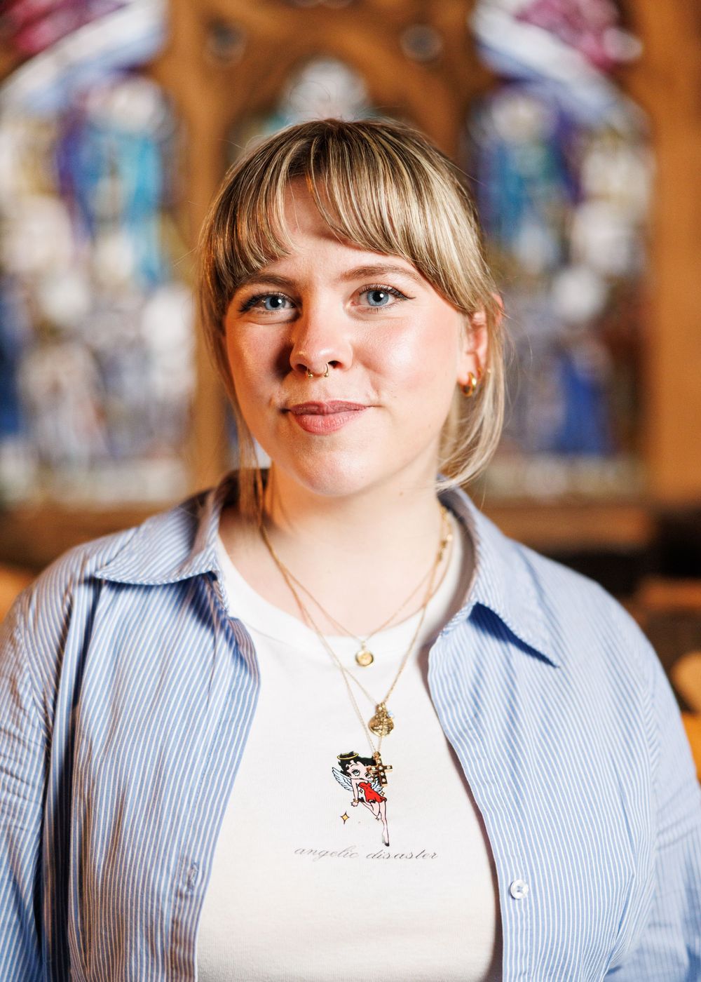 Young woman with blonde bangs smiling in front of colorful stained glass windows in a church.