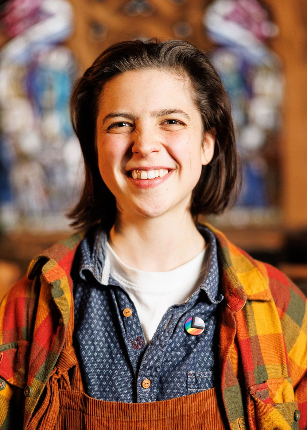 Smiling young person with short dark hair wearing a plaid flannel shirt and overalls, standing in front of a stained glass window.