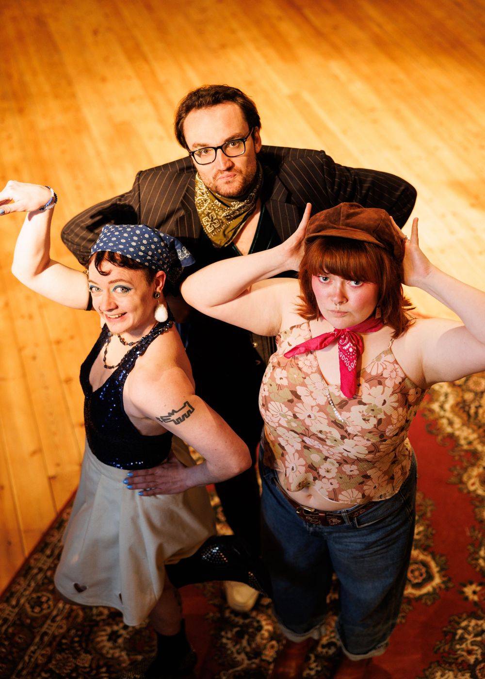Three performers pose dramatically on a wooden floor, two women in retro outfits flanking a man in a pinstripe suit.