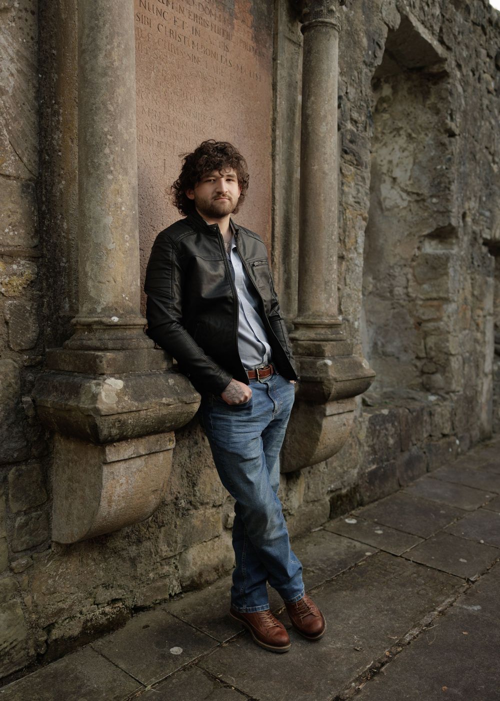 A curly-haired man in a leather jacket and jeans leans against an ancient stone wall with columns.